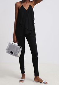Sort lagd sleeveless top, slanke sorte bukser, sølvfarvede flade sandaler og metallic håndtaske. Glat stof, flydende design og minimalistiske accenter.