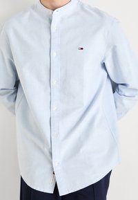 Ljusblå skjorta med knappar och rund krage, fem knappar fram, samt en liten broderad logotyp på vänster bröst.