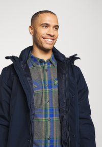 Parka impermeable en color navy con capucha desmontable, llevada sobre una camisa de franela en cuadros grises y azules que cuenta con dos bolsillos en el pecho.