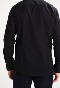 Camisa negra de manga larga hecha de tela suave, con dobladillo curvado, puños con botones y un diseño de cuello clásico.