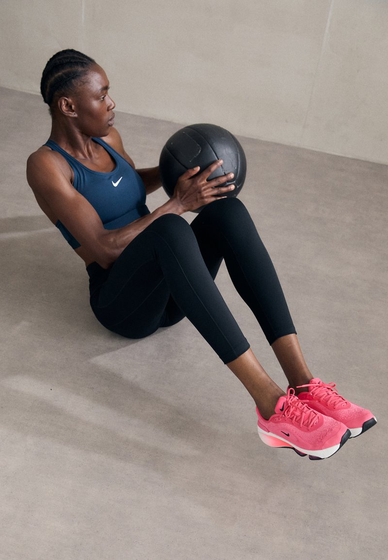 Leggings noirs, brassière de sport bleu marine et chaussures de sport roses sur une personne faisant des redressements assis tout en tenant un ballon médicinal noir sur un fond neutre.
