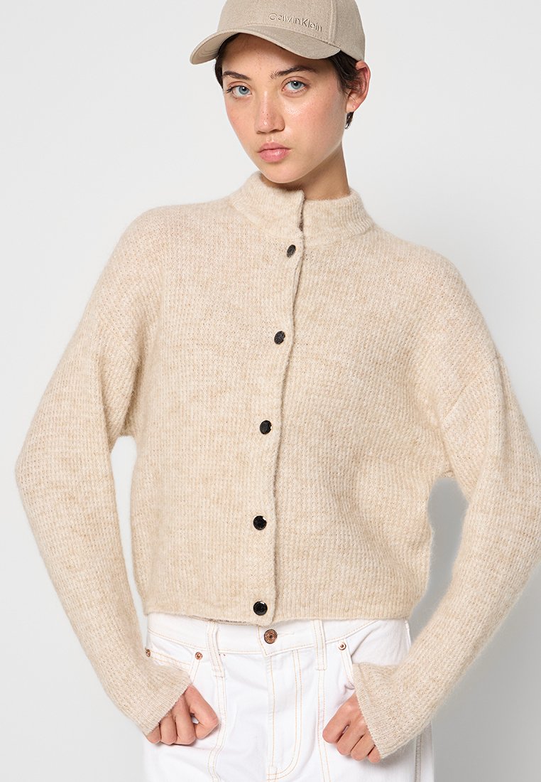Beige Strickjacke mit hohem Kragen, durchgehender Knopfleiste und schwarzen Knöpfen. Weicher, strukturierter Stoff mit langen Ärmeln.