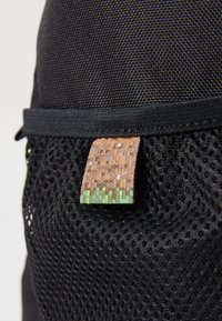 Tessuto a rete nero con un motivo testurizzato, caratterizzato da un'etichetta cucita che mostra un design pixelato marrone e verde.