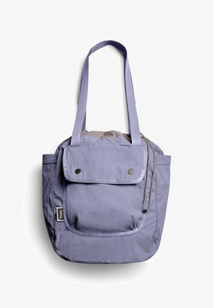 Sac fourre-tout violet clair avec longues anses, poche frontale à rabat sécurisée par deux boutons-pression, et poches latérales sur fond blanc.