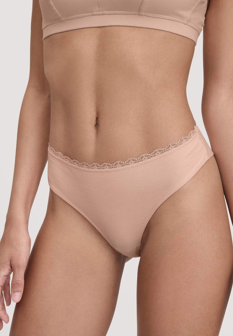 Femme portant des culottes taille haute de couleur chair avec une dentelle et un soutien-gorge assorti de couleur chair sur un fond uni.