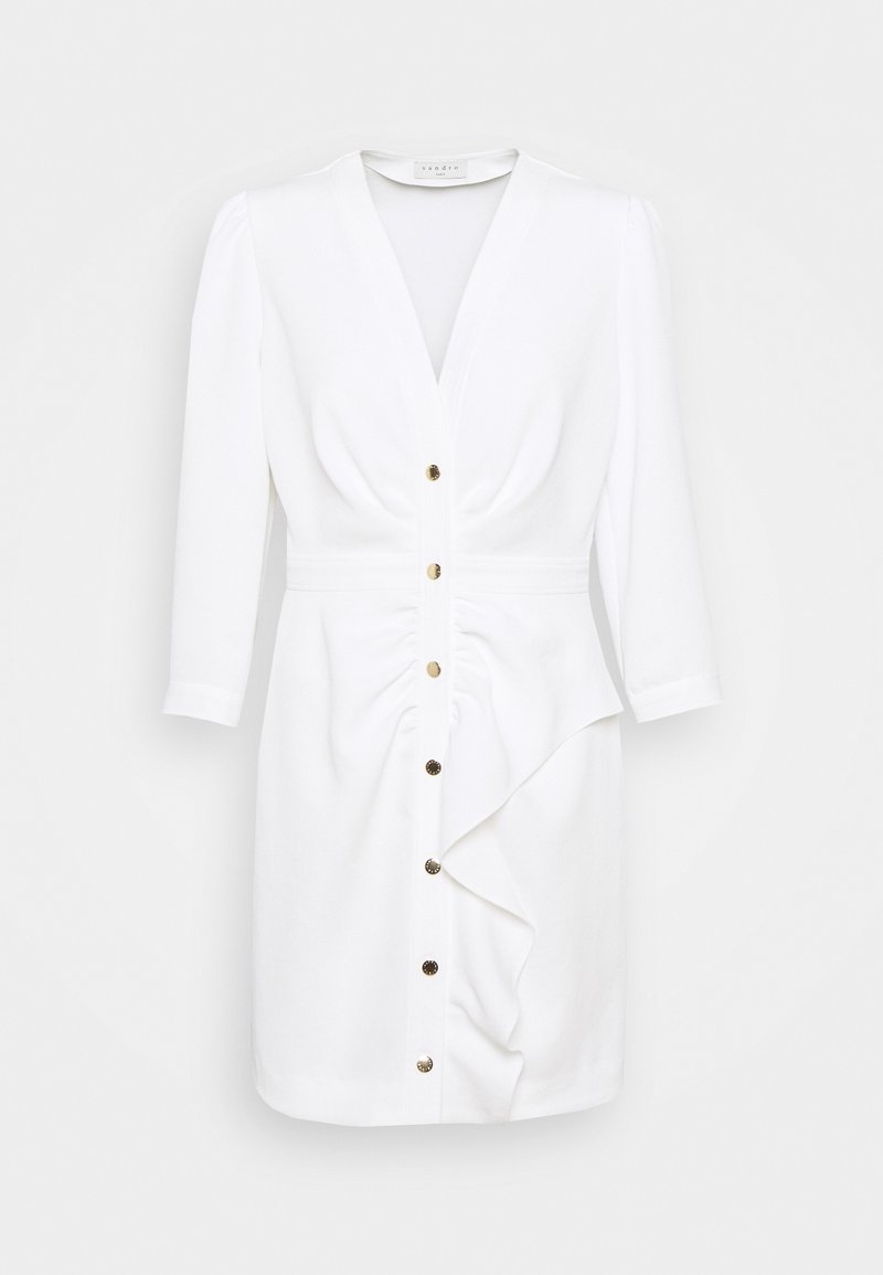 Robe blanche longueur genou avec col en V, manches longues, boutons dorés sur le devant et détail volant asymétrique côté droit.