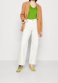 Beige blazer, groen tanktop, witte wijde broekspijpen en groene slides met transparante blokhakken. De outfit combineert een mix van felle kleuren en verschillende texturen.