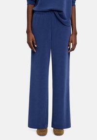 Spodnie o szerokich nogawkach w granatowym materiale, z elastycznym paskiem w talii i gładką fakturą, zestawione z brązowym obuwiem.
