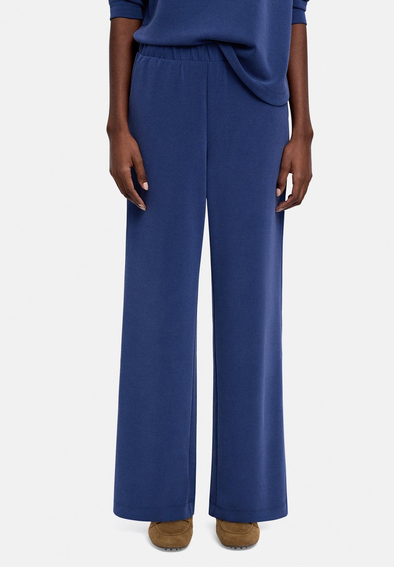 Spodnie o szerokich nogawkach w granatowym materiale, z elastycznym paskiem w talii i gładką fakturą, zestawione z brązowym obuwiem.