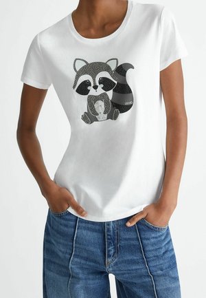 Osoba ubrana w białą koszulkę z krótkim rękawem, z grafiką czarno-szarego szopa ozdobioną małymi kryształkami, w połączeniu z niebieskimi dżinsami.