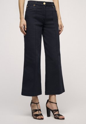 Pantaloni a gamba larga di un blu scuro, realizzati in cotone misto, con tasche anteriori e dettagli con bottoni dorati. Abbinati a sandali neri con tacco alto e cinturini.