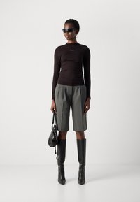 Black long-sleeve top with logo, gray pleated shorts, high black boots, and black handbag; model wearing sleek sunglasses and earrings.