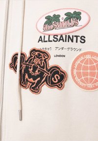 AllSaints beigefärgat tyg med broderade logotyper som inkluderar palmer, en tiger som håller en skivstång, japansk text och en patch med jordklotets design.
