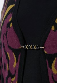 Schwarze Strickjacke mit einem lila und gelben Blumenmuster. Geschlossen mit einer goldfarbenen Brosche, die mit schwarzen Perlen und klaren Akzenten verziert ist.