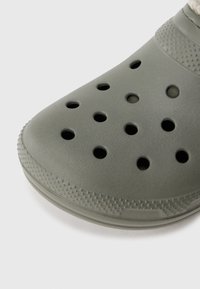 Nahaufnahme eines grauen Gummischuhs mit runden Belüftungslöchern und strukturierter Sohle auf einem schlichten weißen Hintergrund.