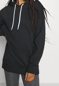 Czarna bluza z kapturem, wykonana z miękkiego materiału, z białymi sznurkami i luźnym krojem, z ściągaczami przy rękawach i u dołu.