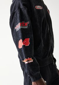 Μαύρο φούτερ με φερμουάρ και υφή υφάσματος. Διαθέτει κόκκινα και λευκά λογότυπα στα μανίκια, συμπεριλαμβανομένου ενός γραφικού με θέμα τους αγώνες και έναν μεγάλο αριθμό «04».