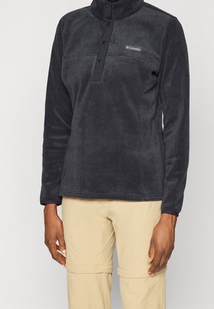 Persona que lleva un polar Columbia negro con botones a presión y pantalones convertibles beige, mostrada desde el cuello hasta mitad del muslo.
