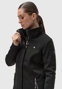 Juoda zip-up striukė su aukštu apykakle, su ribotu tekstūru, šoninėmis užtrauktukų kišenėmis ir sidabrinėmis detalėmis. Viršutiniame kairiajame krūtinės kampe yra logotipas.