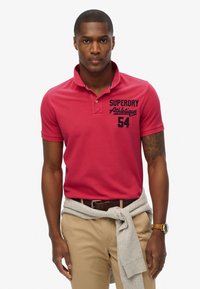 Rød polo t-shirt med krave, med blå logo tryk og nummer. Bæres med beige bukser og en grå sweater bundet om taljen.