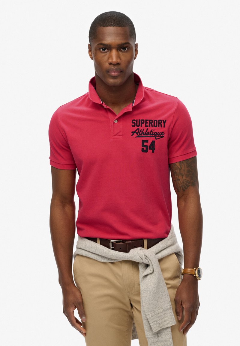 Rød polo t-shirt med krave, med blå logo tryk og nummer. Bæres med beige bukser og en grå sweater bundet om taljen.