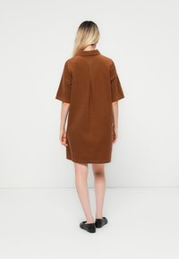 Femme aux longs cheveux blonds portant une robe courte à manches courtes marron et des chaussures noires plates, se tenant de dos contre un fond blanc.