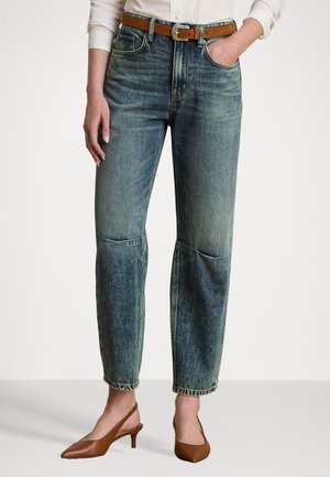 Vrouw draagt vervaagde blauwe hoge taille jeans met kniezemen, een bruine riem, bruine puntige slingback-hakken en een wit overhemd met knopen.