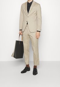 Completo beige su misura con un bottone, camicia nera, scarpe eleganti scure e una borsa tote nera strutturata con manici marroni.