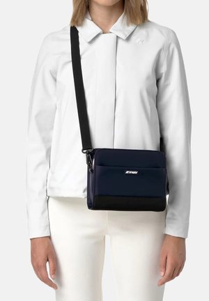 Donna che indossa un completo bianco con una borsa a tracolla K-Way blu navy con tracolla nera regolabile e tasca frontale.