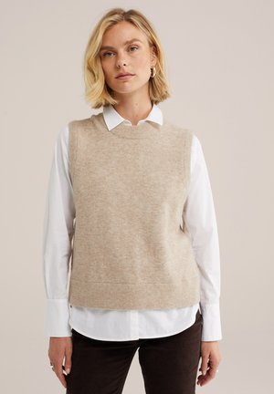 Femme aux cheveux blonds porte un gilet en tricot beige sans manches par-dessus une chemise blanche boutonnée et un pantalon foncé sur un fond beige uni.