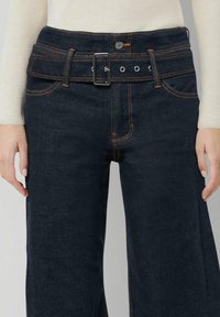 Denimowe jeansy w ciemnoniebieskim kolorze o średnim stanie, z pasującym paskiem z materiału zakończonym metalową klamrą, noszone z jasną bluzką z długim rękawem na szarym tle.