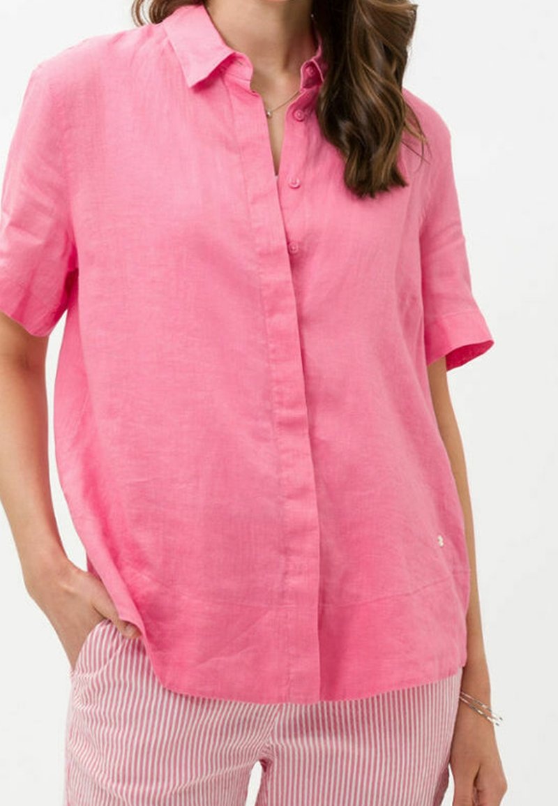 Donna che indossa una camicia a maniche corte rosa brillante con bottoni e pantaloni a righe verticali rosa e bianchi, con una mano in tasca.