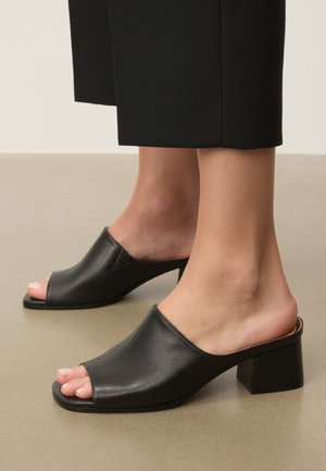 Noge v črnih usnjenih muleih z odprtim prstom in debelimi petami, kombinirane s črnimi skrajšanimi hlačami, stojijo na gladki površini.