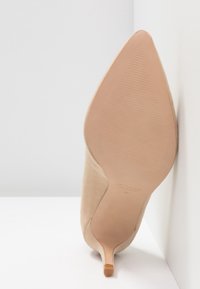 Chaussure à talon haut en daim beige avec un bout pointu et un talon aiguille fin. La semelle est lisse avec des détails de marque gravés.