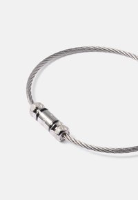 Boucle en câble métallique argenté torsadé avec un fermoir à vis cylindrique sur fond blanc.