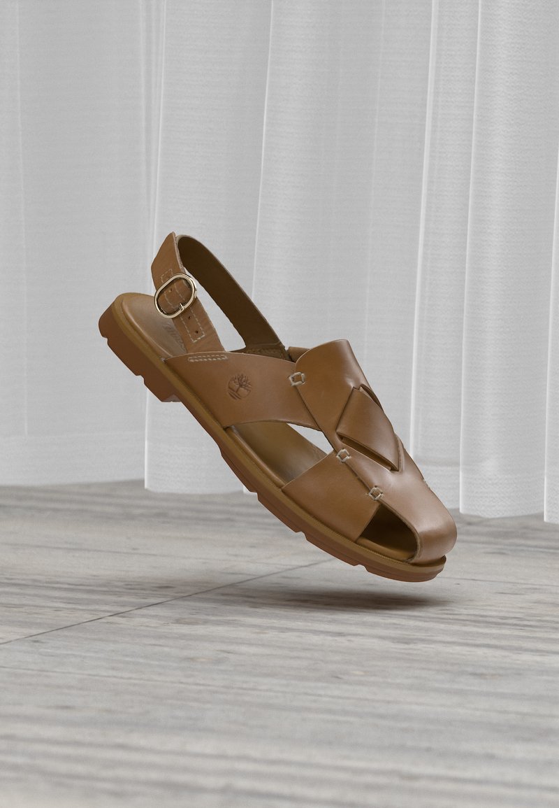 Καφέ δερμάτινο ανδρικό σανδάλι με ρυθμιζόμενο λουράκι με πόρπη και ανοιχτή μύτη, που «αιωρείται» πάνω από ξύλινο δάπεδο ανοιχτού χρώματος, μπροστά από διάφανα λευκά κουρτίνα.