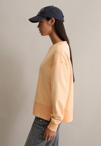 Sweat-shirt couleur pêche avec un col rond et un ourlet côtelé. Assorti à une casquette de baseball bleu marine ornée d'un logo blanc.