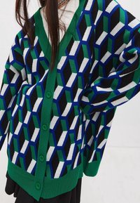 Kvinna som bär en knäpp cardigan med gröna, blåa, svarta och vita geometriska mönster över en vit polotröja.