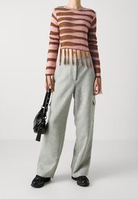 Gestreepte cropped sweater in roze en bruin met franjes aan de onderkant; gecombineerd met grijze wijde broek. Het model houdt een zwarte handtas vast en draagt donkere sneakers.