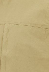 Hellbeige Jacke mit glatter Textur, sichtbaren Nähten und einem schlichten Design. Enthält eine Rückenpasse für Stabilität.