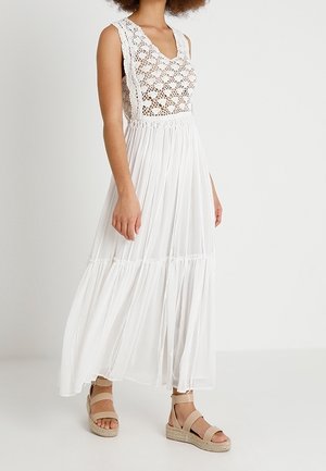 Kobieta w białej, bezrękawnikowej sukience maxi z koronkowym bodice z szydełkową koronką i plisowaną spódnicą, zestawiona z beżowymi sandałami na platformie.