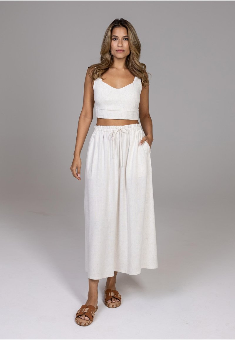 Femme debout en crop top blanc sans manches et pantalon large à cordon, portant des sandales marron sans bride, sur fond gris.