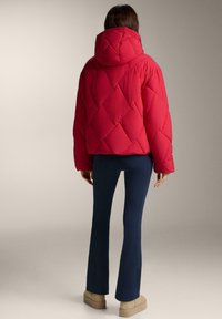 Rød dunjakke med diamantquiltmønster og hætte. Bæres med marineblå bukser og beige fodtøj. Har lange ærmer og en kort pasform.
