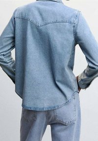 Camicia in denim blu chiaro con design a yoke appuntito, maniche lunghe e orlo curvo; presenta cuciture tonali e polsini con bottoni.