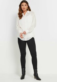 Camicia bianca a maniche lunghe con bottoni e tasca, abbinata a jeans neri attillati e stivaletti neri con tacco. Tessuto liscio, vestibilità su misura.