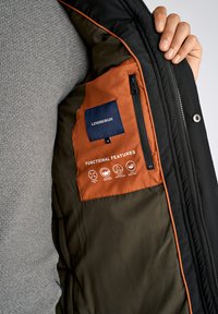 Mustat takki, jossa on oranssi vuori ja tasku, jossa on teksti "LINDBERGH." Ikonit osoittavat ominaisuuksia: mukautuva malli, konepestävä, vedenpitävä ja vettä hylkivä.