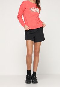 Person klädd i en korallfärgad långärmad The North Face-tröja, svarta shorts, svarta strumpor och svarta sportskor, stående mot en enfärgad bakgrund.