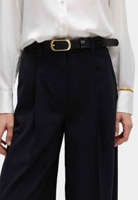 Cinturón de cuero negro con hebilla ovalada dorada, usado sobre pantalones oscuros, combinado con una camisa de satén blanca de botones.