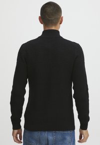 Pull en maille noir avec un col montant, un corps texturé, des poignets en côtes et un ourlet en côtes. Fini lisse avec un motif subtil sur les épaules et le haut du dos.