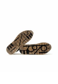 Suole di sneaker con una superficie in gomma textrurata color tan, caratterizzate da un testo nero bold e un design circolare. Costruzione robusta e funzionale.